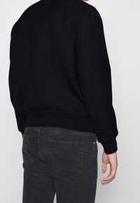 Felpa nera con polsini e orlo a costine; texture liscia. Indossata con jeans grigio scuro caratterizzati da tasche a pattina e un leggero effetto sbiadito.