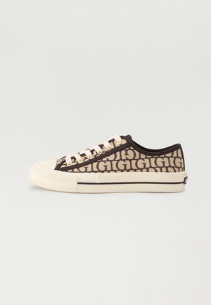 Zapatilla baja de color beige con ribete negro, cordones blancos y patrón repetido "GIGI" en la parte superior de tela, mostrada sobre un fondo liso.