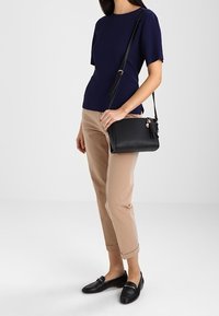 Borsa a tracolla nera con dettaglio a frange, forma rettangolare, texture liscia. Abbinata a una maglia blu navy e pantaloni cropped beige.