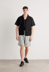 Camisa negra de manga corta con un patrón texturizado, combinada con pantalones cortos grises con estampado de logo. El modelo lleva zapatillas negras. Diseño simple y moderno.