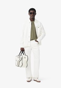 Giacca bianca in cotone sopra una camicia verde oliva, abbinata a pantaloni bianchi e sneakers beige chiaro. Tieni in mano una borsa bianca con dettagli neri e logo.