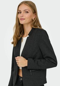 Černý přizpůsobený saka s texturovaným povrchem, který má minimalistický design, kapsy na zip a klopový límec. V kombinaci s přiléhavým bílým topem.