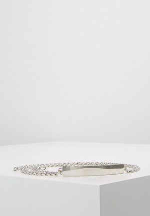 Collier chaîne en argent avec une plaque d'identification rectangulaire polie, posée à plat sur une surface blanche inclinée.