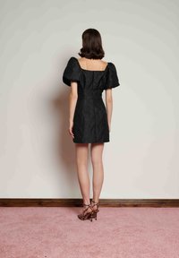 Černé krátké šaty s nafouklými rukávy, texturovanou látkou, čtvercovým výstřihem na zádech a přiléhavým pasem. Kombinované s páskovými botami na vysokém podpatku.