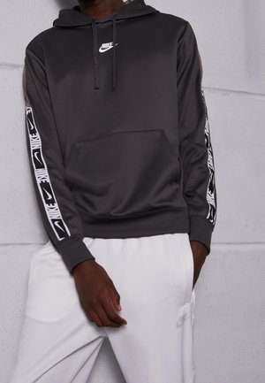 Mężczyzna ubrany w czarną bluzę z kapturem Nike z paskami z logo na rękawach oraz białe spodnie dresowe Nike, stojący na tle jasnoszarej ściany.