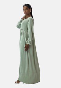 Langes, fließendes Kleid aus hellgrünem Stoff mit langen  Ärmeln und geraffter Taille, ausgestattet mit rundem Ausschnitt und weicher Textur.