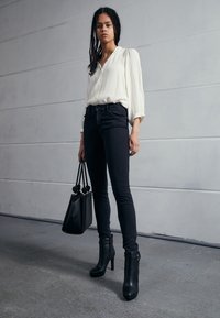 Cream blouse with puffed sleeves, dark skinny jeans, black heeled ankle boots, and a black textured handbag. Minimalist urban setting.