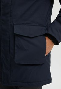 Veste bleu marine avec une finition lisse et mate. Présente une poche plaquée sur le côté, fabriquée en tissu durable avec des coutures visibles.