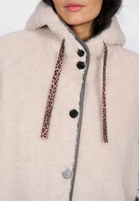 Kremowy płaszcz z futra sztucznego z kapturem, z kontrastowym szarym wykończeniem, sznurkami w panterkę oraz czarnymi guzikami. Miękka tekstura i przytulny design.