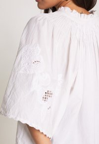 Blusa bianca con maniche corte e ampie, caratterizzata da ricami floreali e dettagli a intaglio. Collo arricciato e tessuto morbido e leggero.