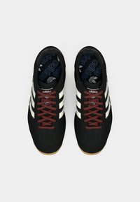 Black athletic shoes with three white stripes, maroon laces, textured materials, and gum rubber soles; branding on the insoles.