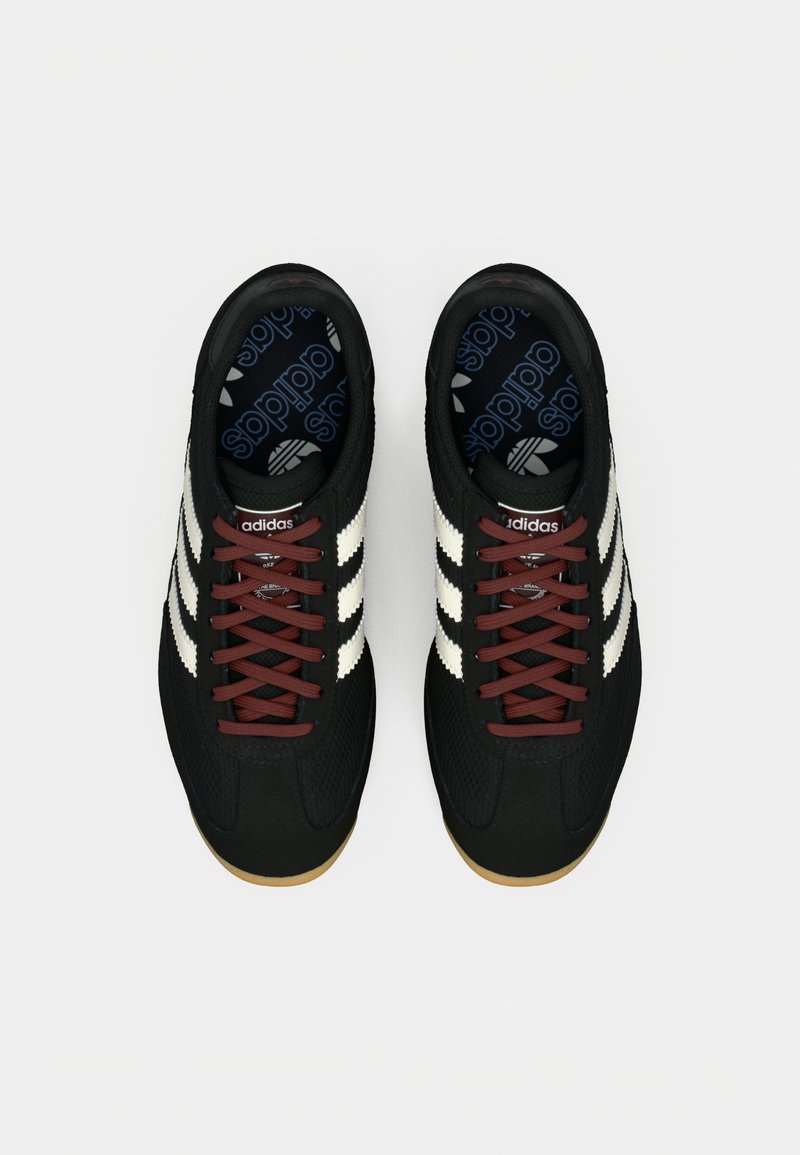 Zapatillas deportivas negras con tres rayas blancas, cordones color burdeos, materiales texturizados y suelas de goma. Marca en las plantillas.