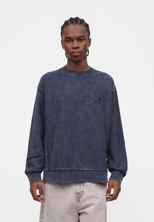 Jonge man met gevlochten haar, een gewassen donkerblauwe trui en lichtbeige broek, staand tegen een effen lichte achtergrond.