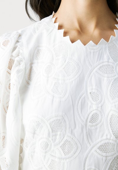 Haut blanc ornés de motifs floraux brodés, avec un décolleté festonné et des manches bouffantes. Le tissu a un aspect doux et texturé.