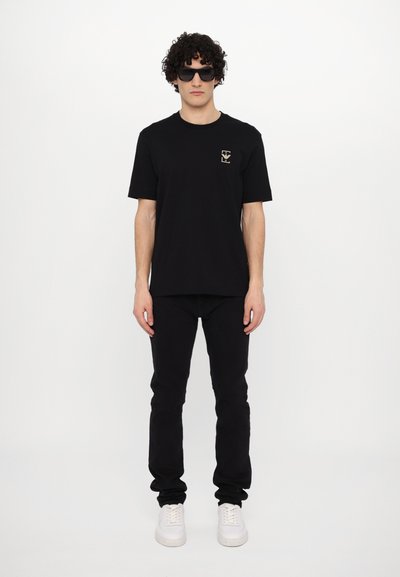 Juoda medvilninė marškinėliai su mažu auksiniu logotipu kairėje krūtinės pusėje, derinami su juodomis džinsomis ir baltomis sportbačiais. Modelis dėvi juodus akinius nuo saulės.