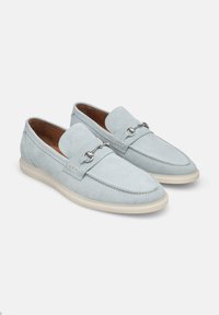 Mocassini in pelle scamosciata blu chiaro con design slip-on, che evidenziano una punta cucita e hardware metallico decorativo sul pannello frontale. Suola in gomma.