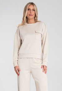 Beżowy sweatshirt z klasycznym okrągłym dekoltem, długimi rękawami i kieszonką naszywaną na lewej piersi. Gładka tekstura, luźny krój.