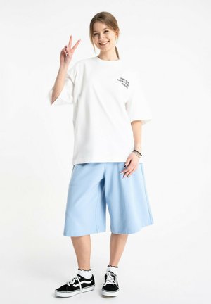 Młoda kobieta uśmiecha się, nosi białą, oversize'ową koszulkę z napisem, jasnoniebieskie szerokie spodenki, czarne trampki, pokazując znak pokoju prawą ręką.