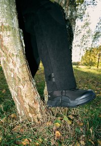 Fekete túracipő, hálós felsőrésszel és gumitalppal, egy fával szemben elhelyezve. A környező területen fű és lehullott levelek találhatók.