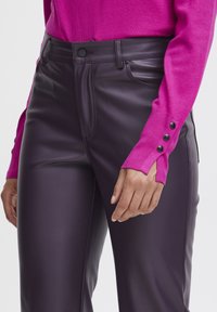Lilla faux læderbukser med en glat tekstur, der har forlommer og en sort knaplukning. Kombineret med en lys pink sweater.