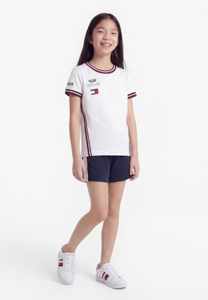 Χαμογελαστή κοπέλα στέκεται, φορώντας λευκό μπλουζάκι με λογότυπο και κόκκινο και μπλε περίγραμμα, σκούρο μπλε σορτς και λευκά αθλητικά παπούτσια με κόκκινες και μπλε ρίγες.