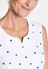 Weißes ärmelloses Kleid mit V-Ausschnitt, versehen mit kleinen schwarzen Blumen- und Blattformen. Strukturiertem Stoff mit dezenten Verzierungen.