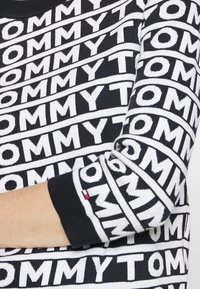 Schwarzer und weißer Strickpullover mit wiederholtem "TOMMY"-Textmuster, gerippten Bündchen und glatter Textur.