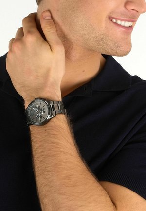 Uomo che indossa una polo nera si tocca il collo con la mano sinistra, mostrando un orologio cronografo argento con cinturino in metallo al polso, sorridendo.