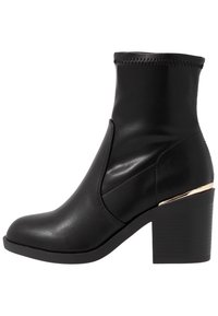 Bottine noire en cuir lisse avec un bout rond, présentant un haut côtelé et un talon bloc rehaussé d'accents dorés avec des lignes horizontales.
