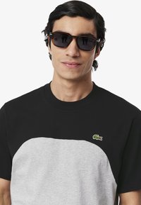 Camiseta de manga corta con una sección superior negra y una sección inferior gris, con un logo de cocodrilo verde en el pecho. Material de algodón texturizado.