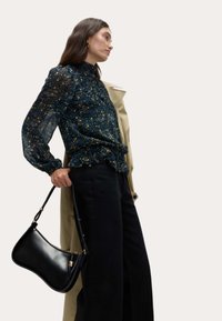 Sort læder skuldertaske med en buet form, guldfarvede detaljer og lynlås. Båret med en bluse med blomsterprint og mørke bukser.