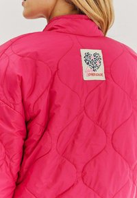 Różowa pikowana kurtka z metką w wytłoczony wzór w panterkę oraz napisem "kochaj inne kolory". Gładka tekstura z falistym wzorem.