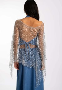 Chal de punto abierto plateado con bordes de flecos, usado sobre un top halter azul y una falda de aspecto vaquero, que muestra un diseño texturizado en capas.