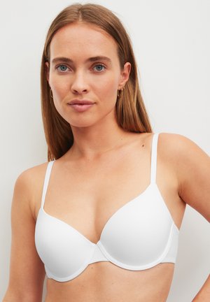 Giovane donna con capelli lisci castano chiaro che indossa un reggiseno bianco modellato con ferretto, su sfondo neutro