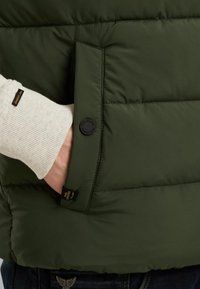 Olive-green puffer vest featuring a side pocket with a circular black button, quilted texture, and branding tag on the hem.