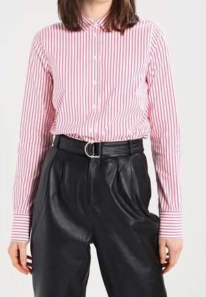 Person iført en rød og hvid lodret striped skjorte med knapper, stukket ned i højtaljede sorte læderbukser med et bælte med dobbelt ring.