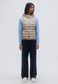 Μπεζ αμάνικο μπουφάν με κουκούλα, διαθέτει κάθετο καπιτονέ σχέδιο, συνδυασμένο με ανοιχτό μπλε μπλούζα μακρυμάνικη, μαύρο παντελόνι φαρδύ και λευκά παπούτσια.