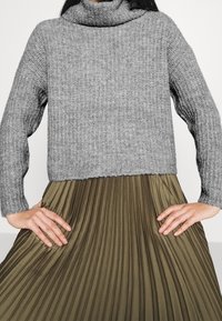 Grauer Strickpullover mit Rollkragen und strukturierter Oberfläche, kombiniert mit einem plissierten olivgrünen Rock, Hände in die Hüften gestützt.