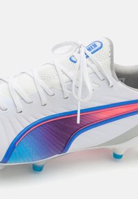 Botín de fútbol blanco con parte superior de malla, franja en degradado de púrpura a rosa, acentos azules y sistema de cordones estándar. Detalle de superficie texturizada.