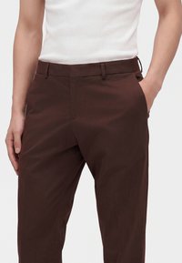 Homme portant un pantalon marron et un haut blanc sans manches, une main dans la poche, sur un fond uni.