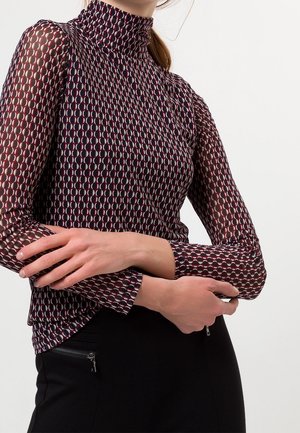 Vrouw draagt een langarmige, patroon coltrui met rode, witte en zwarte cirkelvormige ontwerpen, armen gekruist over zwarte broek.