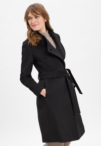 Zwart wollen jas met een ceintuur in de taille, grote kraag en schuine zijzakken. Gladde textuur en getailleerde pasvorm.