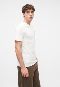 Νεαρός άντρας με καστανά σγουρά μαλλιά που φοράει λευκό κοντομάνικο μπλουζάκι και καφέ παντελόνι, στέκεται μπροστά σε ένα απλό λευκό φόντο.