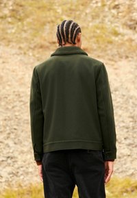 Person with braided hair wearing a dark green jacket and black pants standing outdoors on a gravel surface facing away.