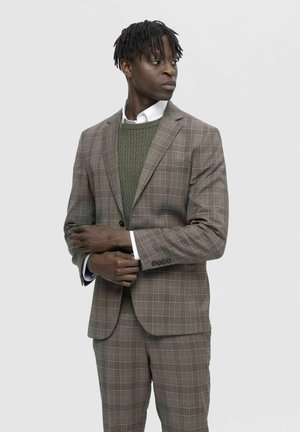 Brown plaid suit with a textured olive green sweater underneath, white collared shirt, and subtle button accents on the sleeves.