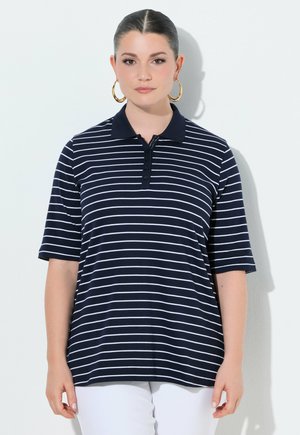 Kvinde med tilbagelagt hår, iført store guldhoop-øreringe og en marineblå polo med hvide vandrette striber, stående foran en hvid baggrund.