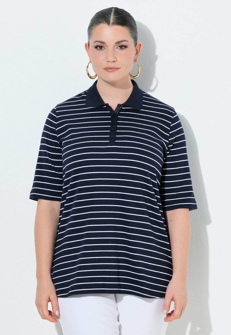 Kobieta z zaczesanymi do tyłu włosami, nosząca duże złote kolczyki koła oraz granatową koszulkę polo z białymi poziomymi paskami, stojąca na białym tle.