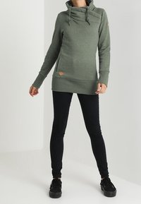 Sudadera larga verde con un alto cuello, cordón ajustable y detalles acanalados laterales; combinada con leggings negros y zapatillas negras.