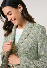Green tweed blazer with a textured pattern, light purple accents, notched lapels, and a two-button closure. Model is adjusting the collar.