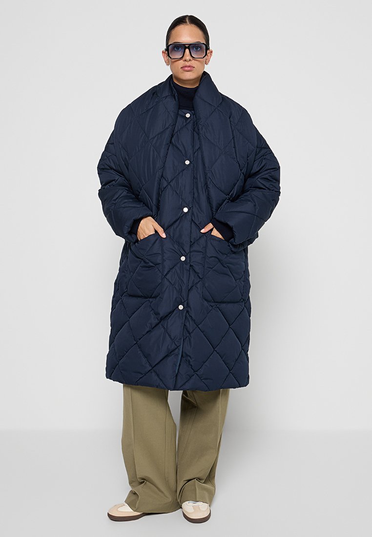 Granatowa pikowana kurtka puchowa z wysokim kołnierzem, zatrzaskami i dużymi kieszeniami, w zestawieniu z oliwkowo-zielonymi spodniami o szerokich nogawkach i beżowymi butami.